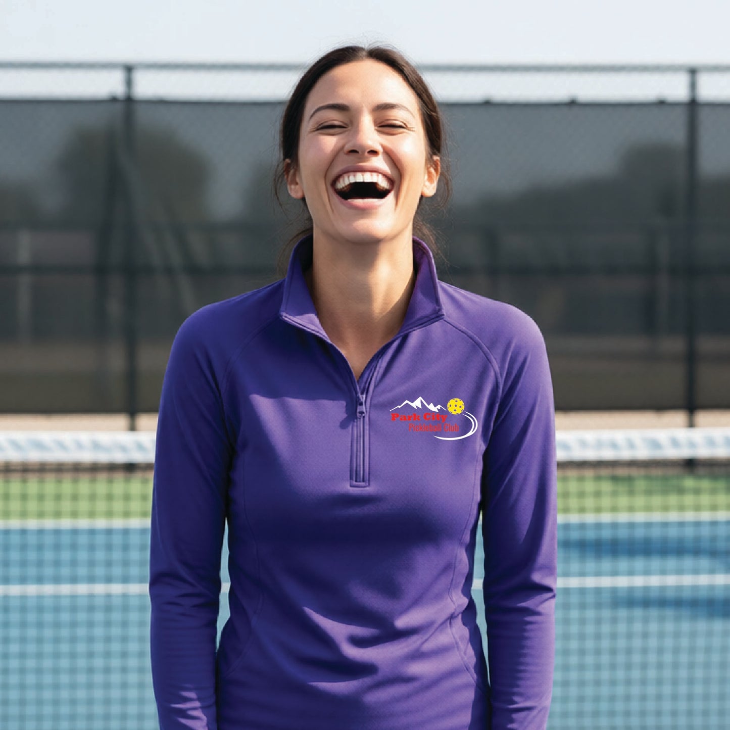 A woman on an outdoor pickleball court, wearing a purple long-sleeve quarter-zip athletic top with a custom 'Park City Pickleball Club' logo printed on the pocket area.