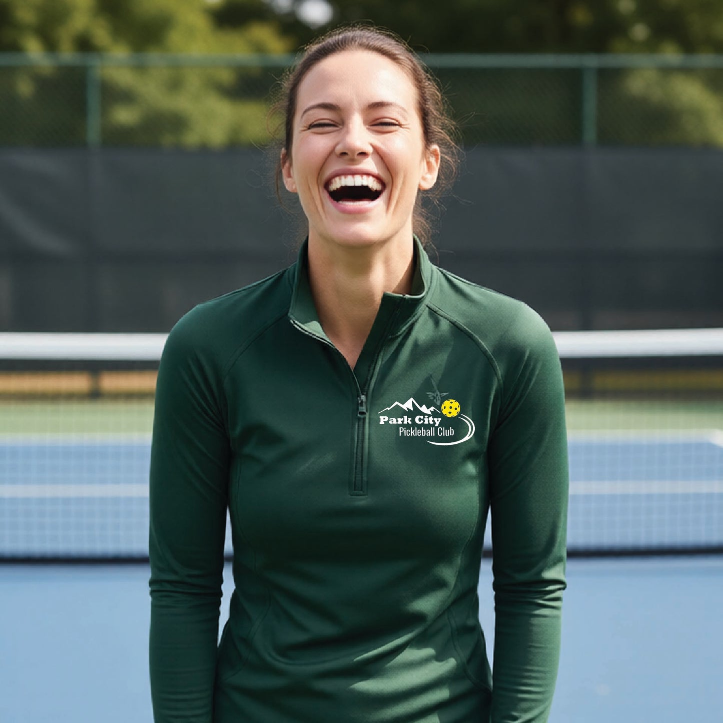 A woman on an outdoor pickleball court, wearing a forests green long-sleeve quarter-zip athletic top with a custom 'Park City Pickleball Club' logo printed on the pocket area.