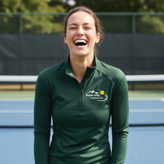 A woman on an outdoor pickleball court, wearing a forests green long-sleeve quarter-zip athletic top with a custom 'Park City Pickleball Club' logo printed on the pocket area.