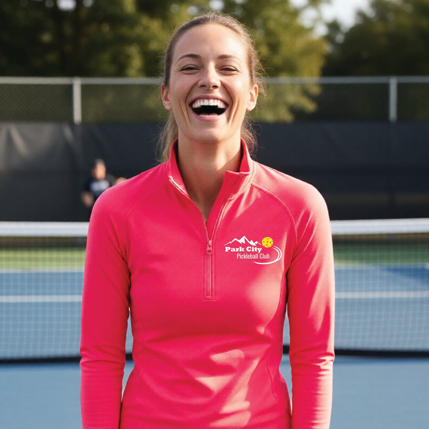 A woman on an outdoor pickleball court, wearing a hot coral pink long-sleeve quarter-zip athletic top with a custom 'Park City Pickleball Club' logo printed on the pocket area.