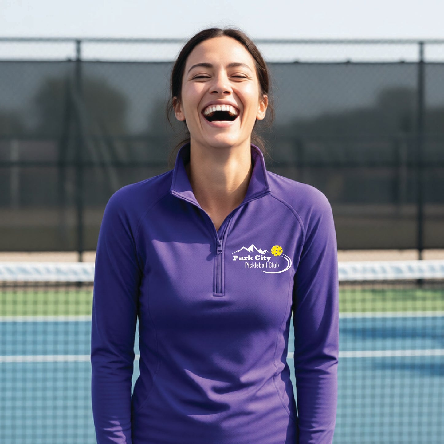 A woman on an outdoor pickleball court, wearing a purple long-sleeve quarter-zip athletic top with a custom 'Park City Pickleball Club' logo printed on the pocket area.