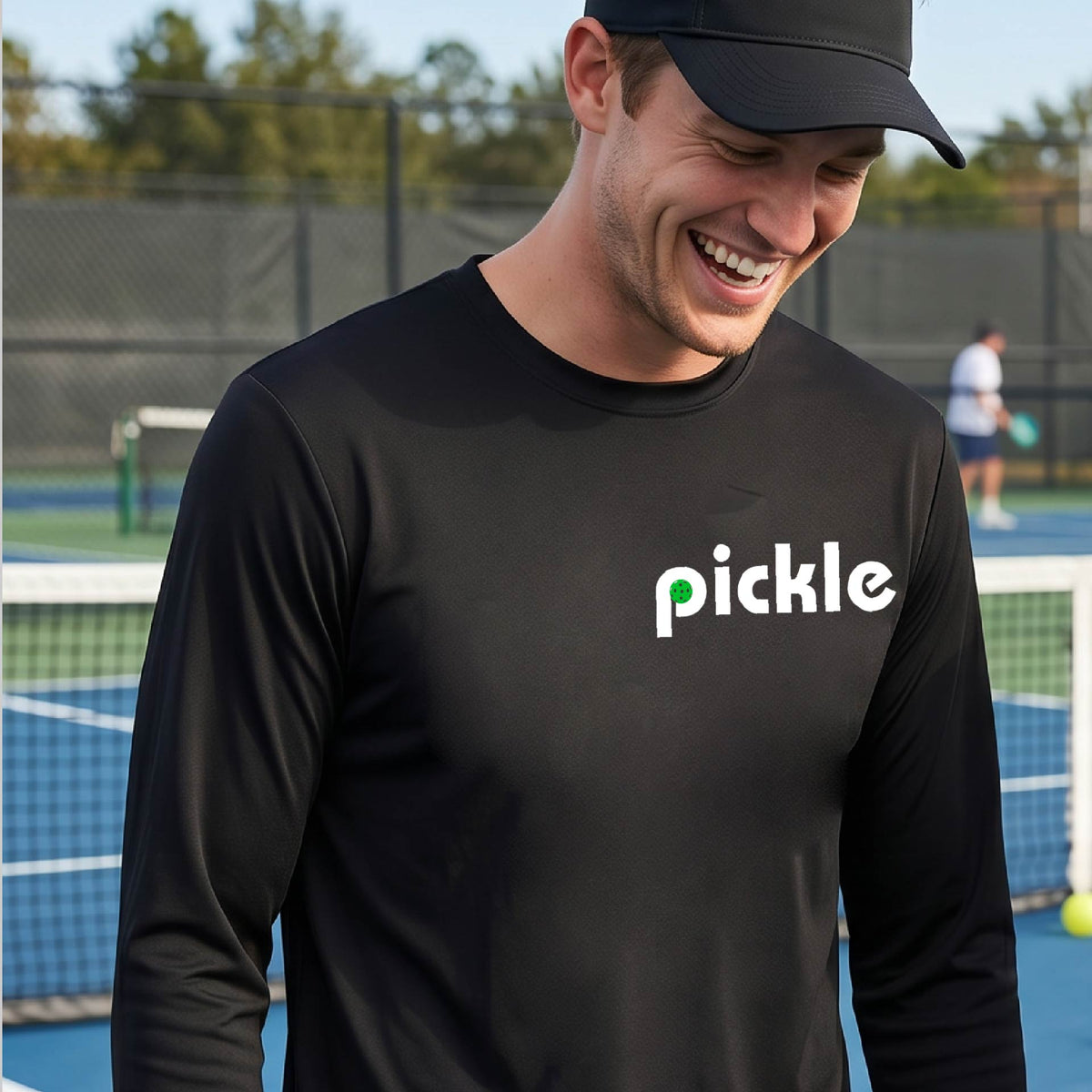 A man wearing a black long-sleeve t-shirt with a white graphic of ‘PICKLE’ with a green pickleball integrated into the letter P printed on the front, in front of a pickleball court. 