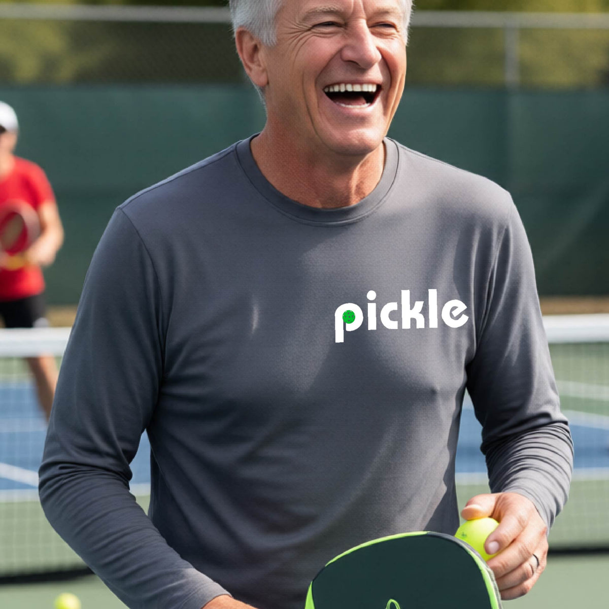 A man wearing a gray long-sleeve t-shirt with a white graphic of ‘PICKLE’ with a green pickleball integrated into the letter P printed on the front, in front of a pickleball court. 