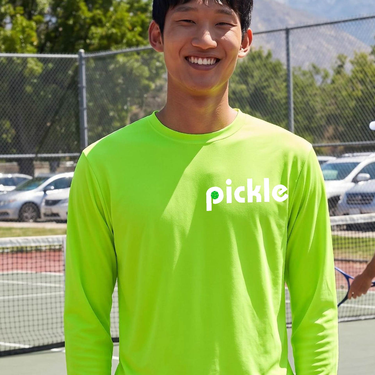A man wearing a lime green long-sleeve t-shirt with a white graphic of ‘PICKLE’ with a green pickleball integrated into the letter P printed on the front, in front of a pickleball court. 