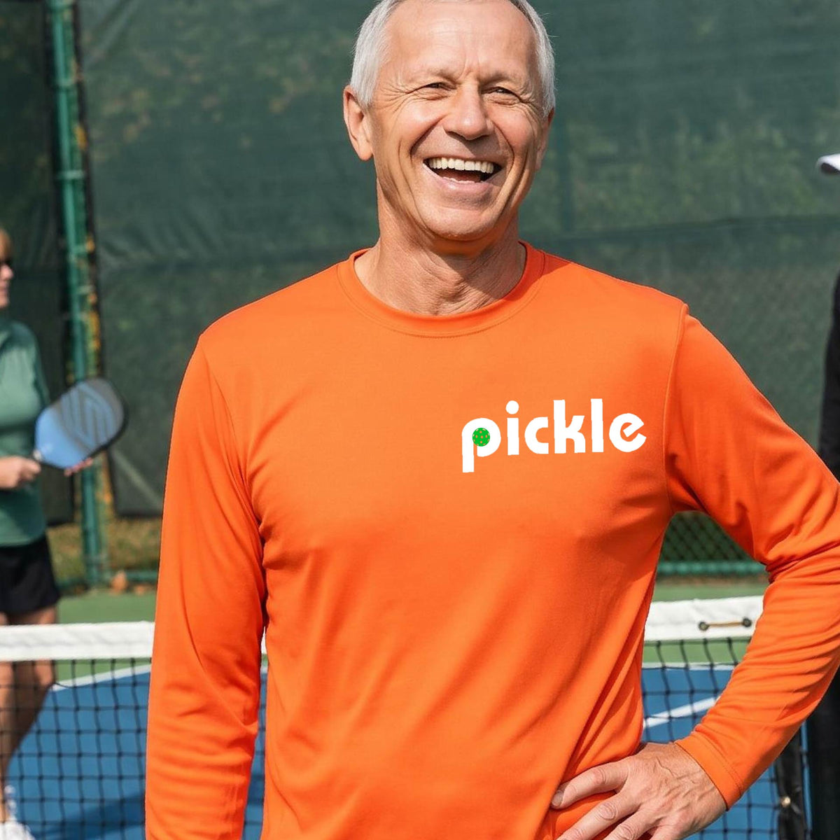 A man wearing an orange long-sleeve t-shirt with a white graphic of ‘PICKLE’ with a green pickleball integrated into the letter P printed on the front, in front of a pickleball court. 