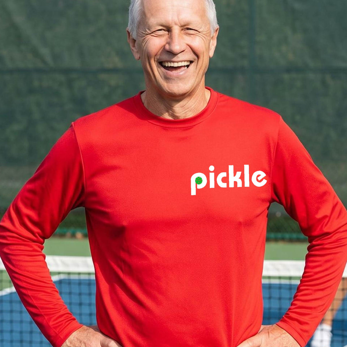 A man wearing a red long-sleeve t-shirt with a white graphic of ‘PICKLE’ with a green pickleball integrated into the letter P printed on the front, in front of a pickleball court. 