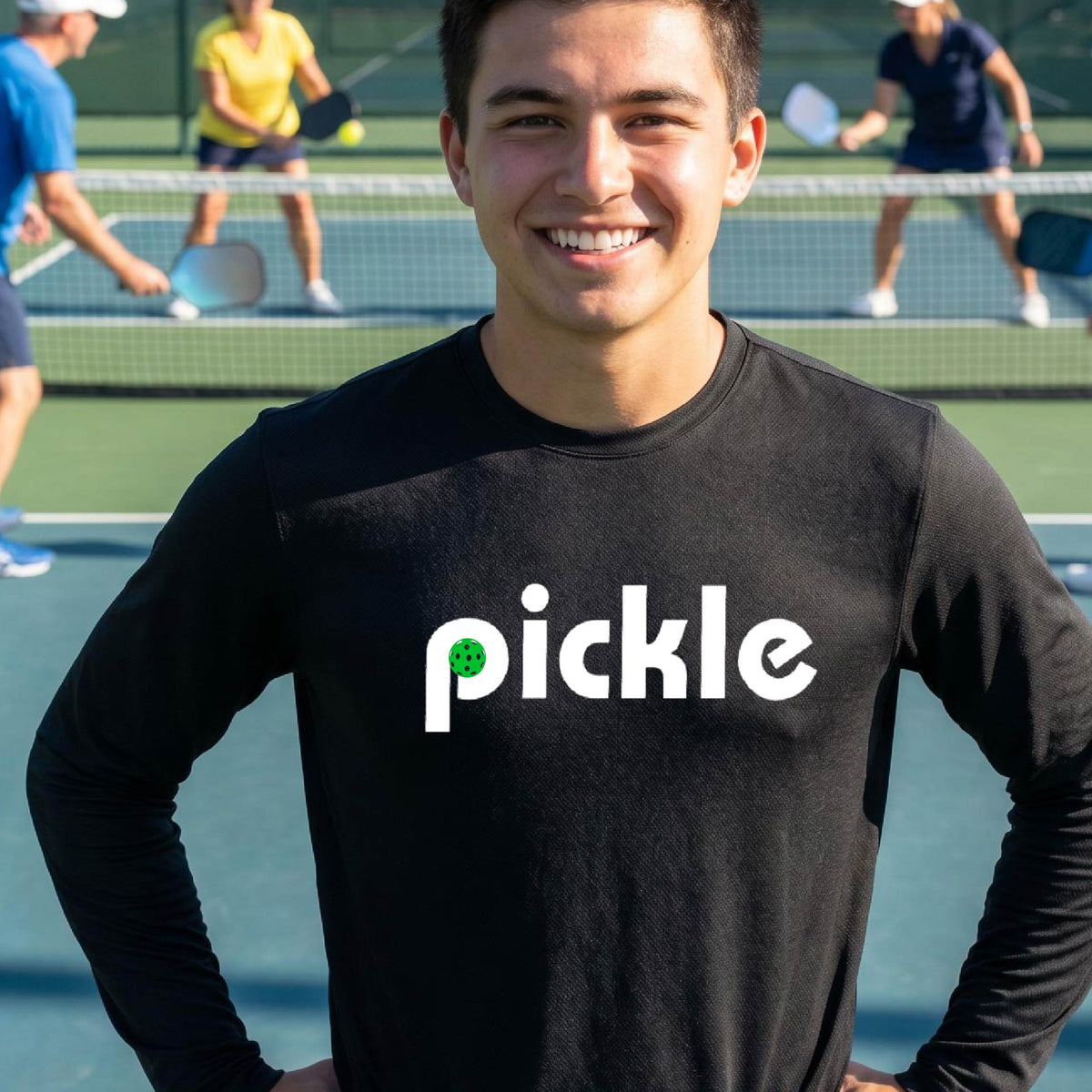 A man wearing a black long-sleeve t-shirt with a white graphic of ‘PICKLE’ with a green pickleball integrated into the letter P printed on the front, in front of a pickleball court. 