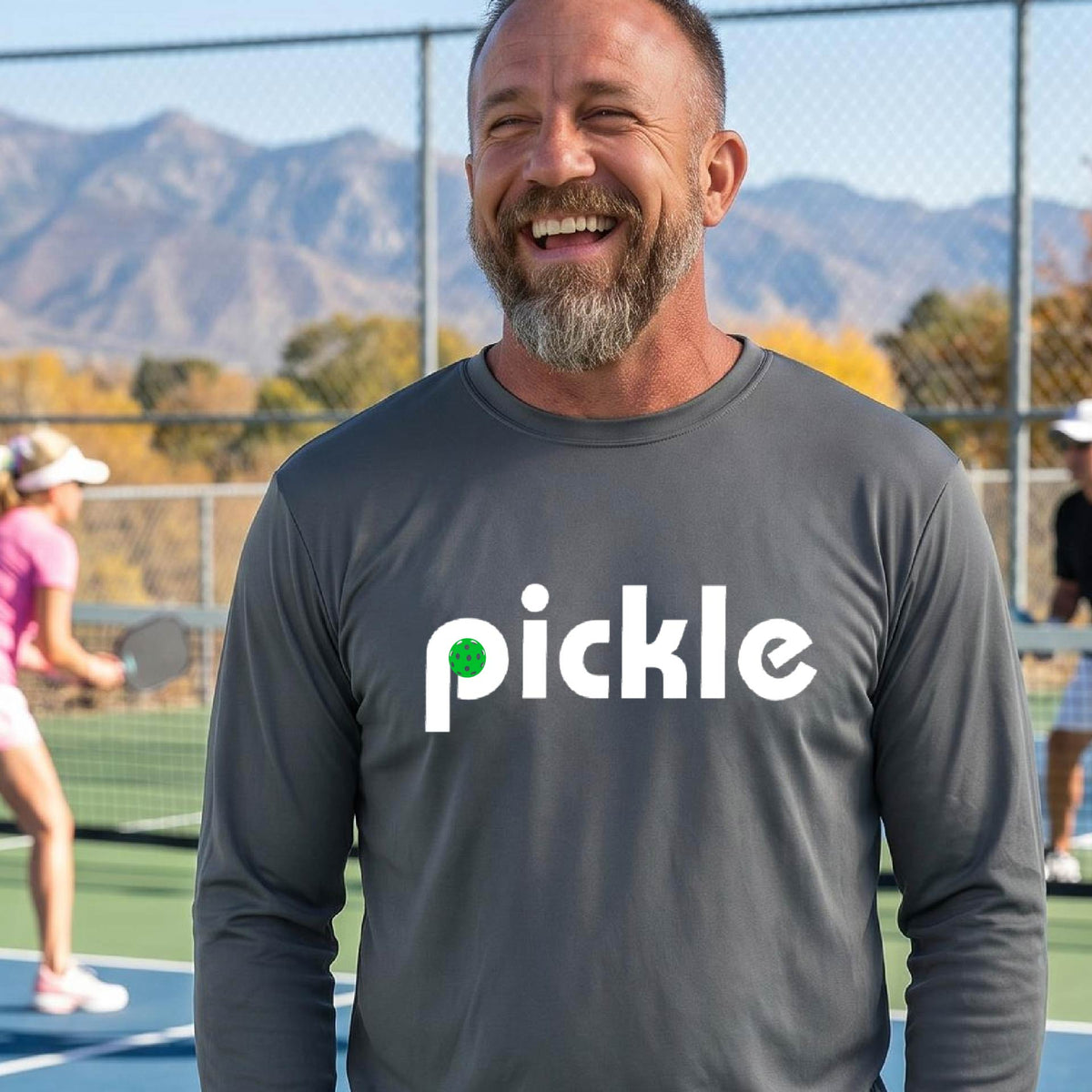 A man wearing a gray long-sleeve t-shirt with a white graphic of ‘PICKLE’ with a green pickleball integrated into the letter P printed on the front, in front of a pickleball court. 