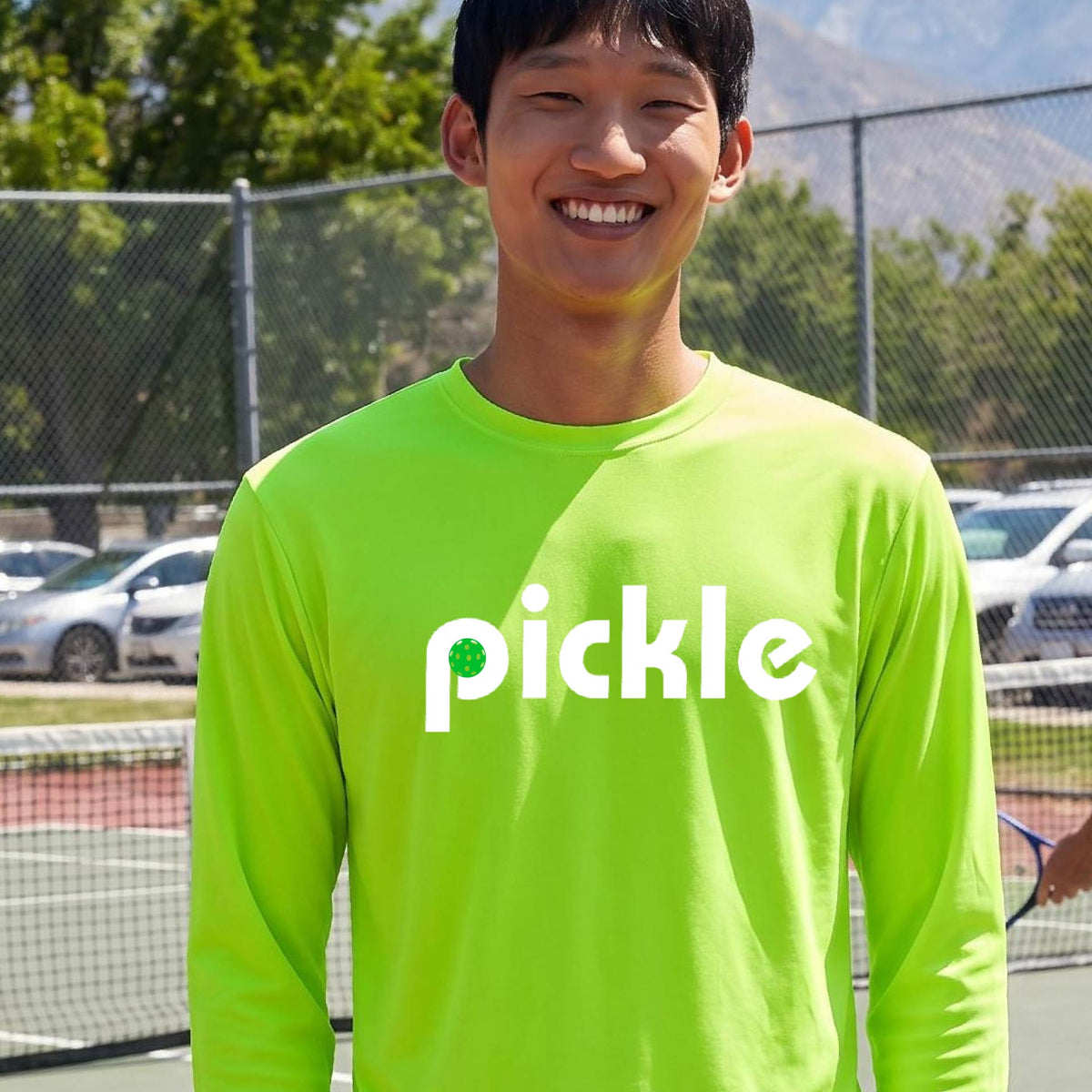 A man wearing a lime green long-sleeve t-shirt with a white graphic of ‘PICKLE’ with a green pickleball integrated into the letter P printed on the front, in front of a pickleball court. 