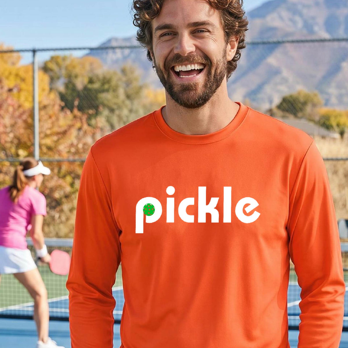 A man wearing an orange long-sleeve t-shirt with a white graphic of ‘PICKLE’ with a green pickleball integrated into the letter P printed on the front, in front of a pickleball court. 