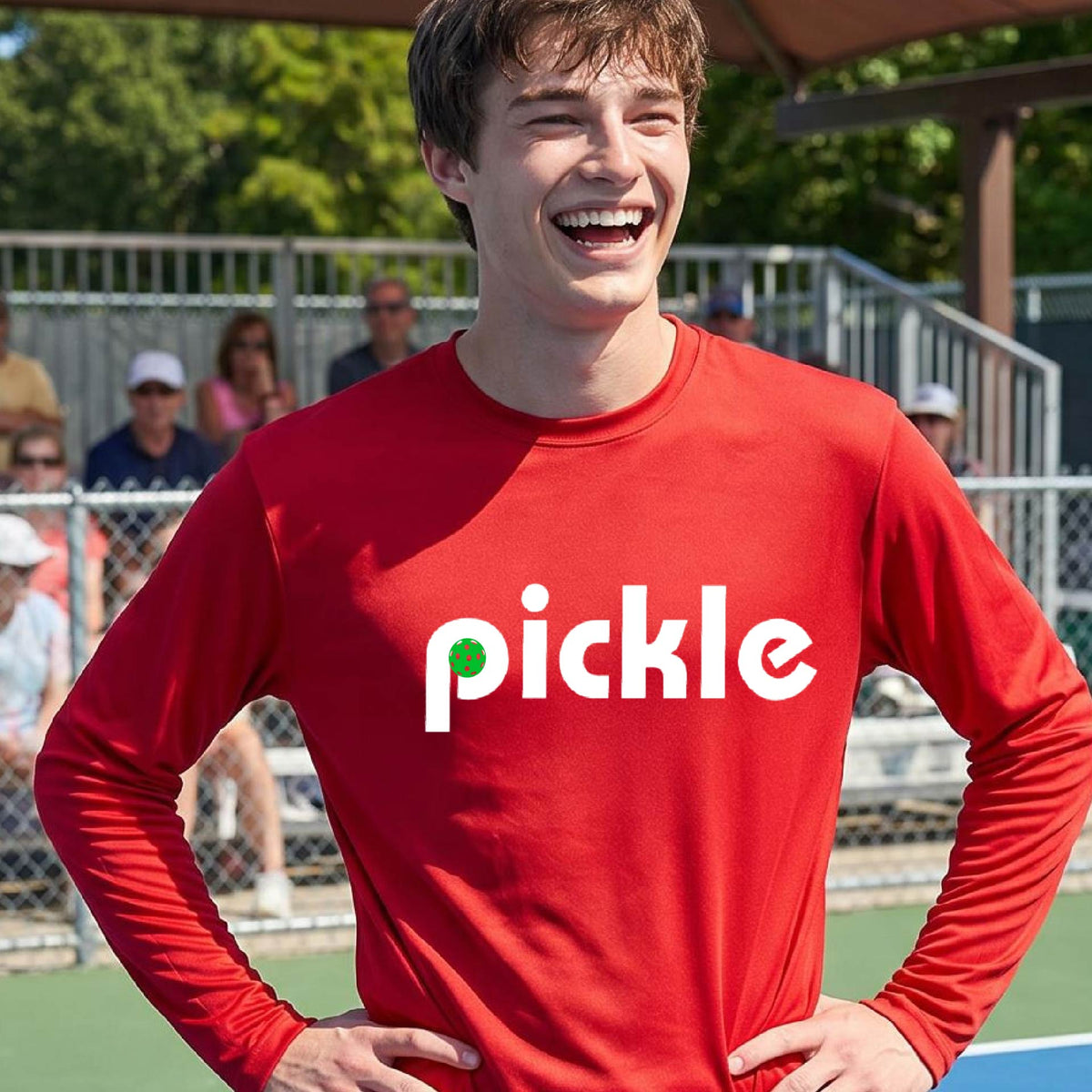 A man wearing a red long-sleeve t-shirt with a white graphic of ‘PICKLE’ with a green pickleball integrated into the letter P printed on the front, in front of a pickleball court. 