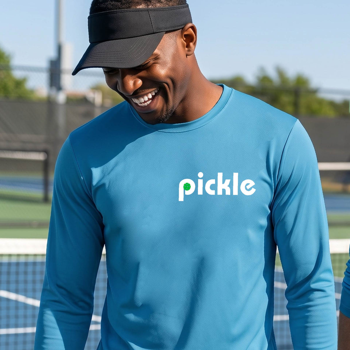 A man wearing a blue long-sleeve t-shirt with a white graphic of ‘PICKLE’ with a green pickleball integrated into the letter P printed on the front, in front of a pickleball court. 