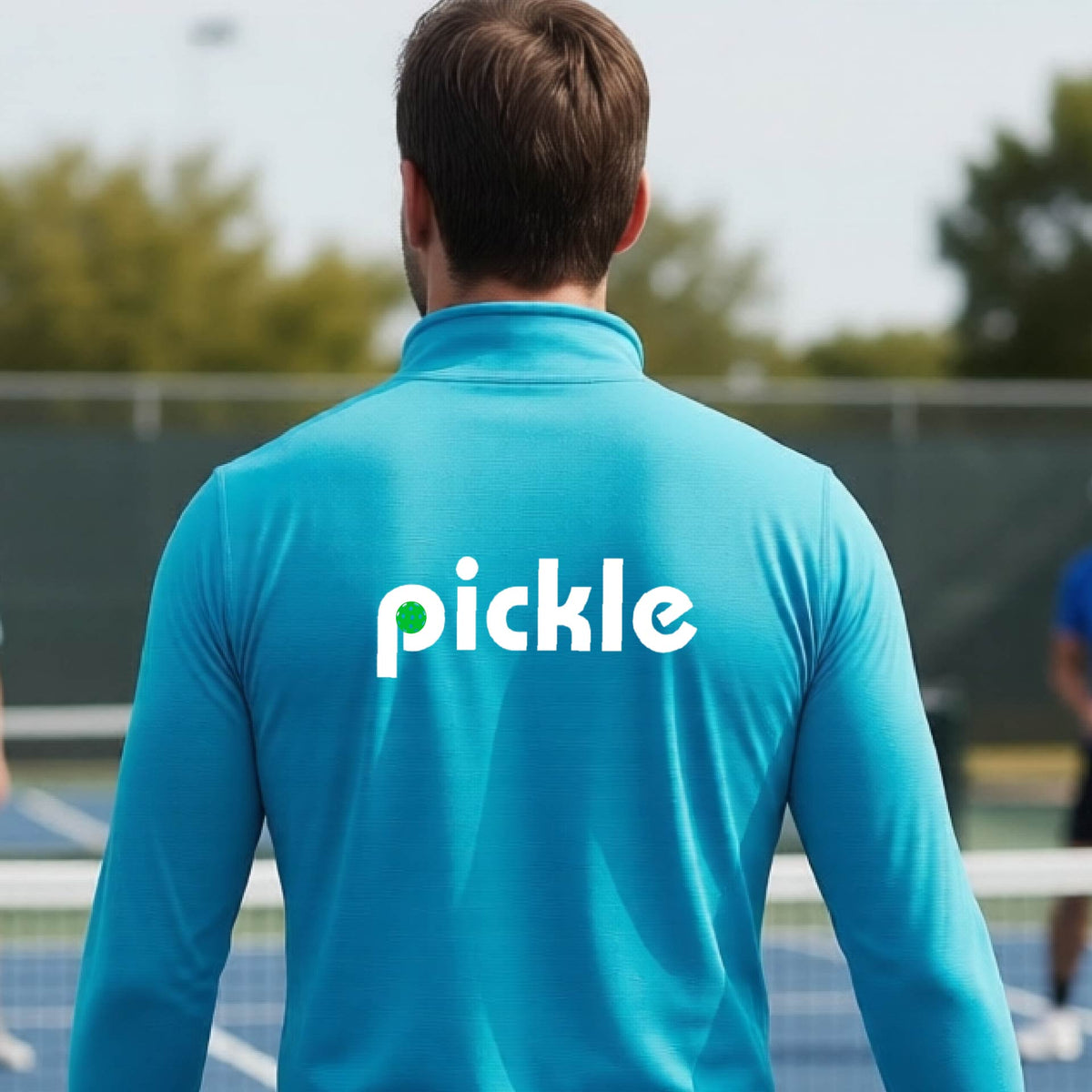 A man wearing a blue quarter-zip shirt with a white graphic of ‘PICKLE’ with a green pickleball integrated into the letter P printed on the back, in front of a pickleball court. 