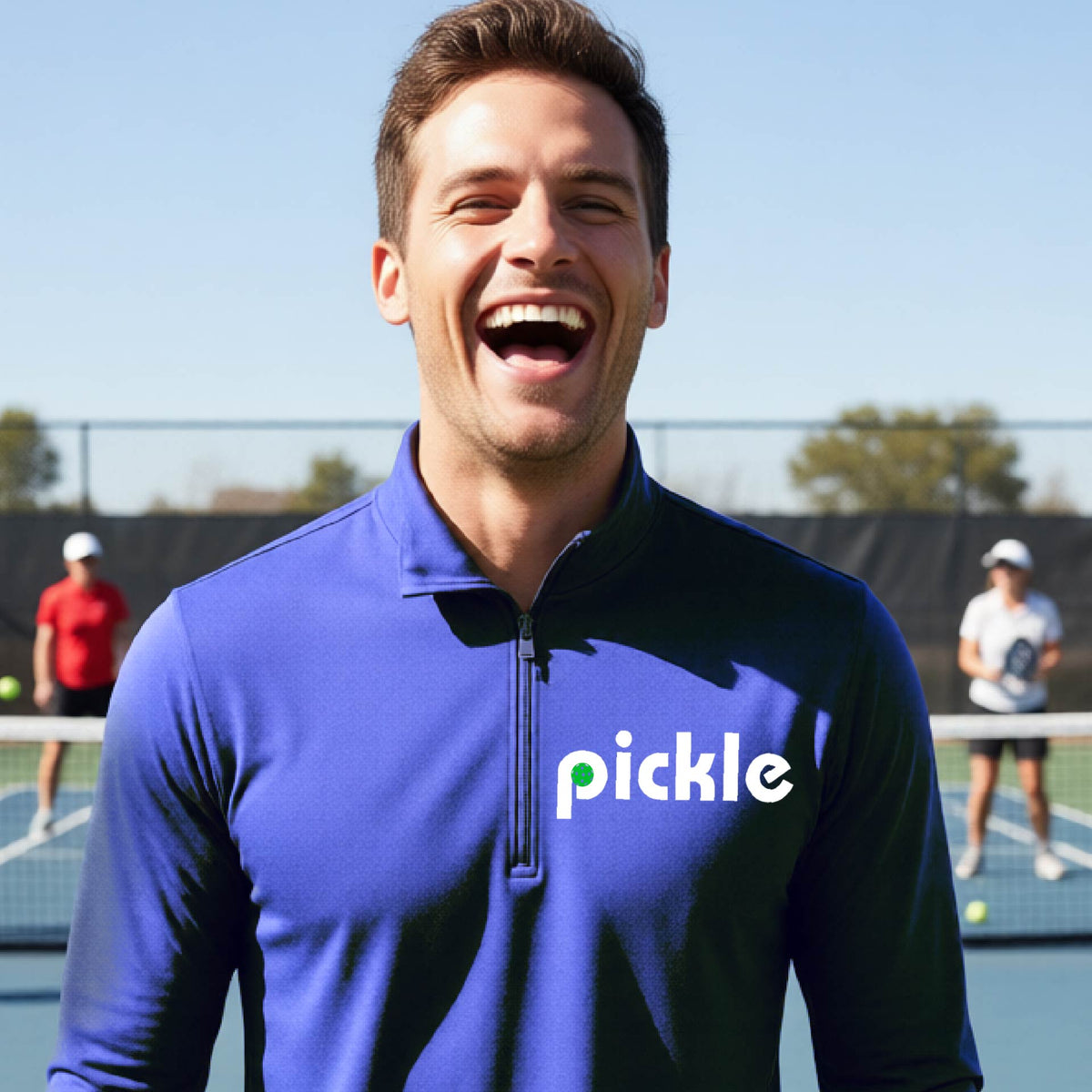 A man wearing a royal blue quarter-zip shirt with a white graphic of ‘PICKLE’ with a green pickleball integrated into the letter P printed on the front in front of a pickleball court. 