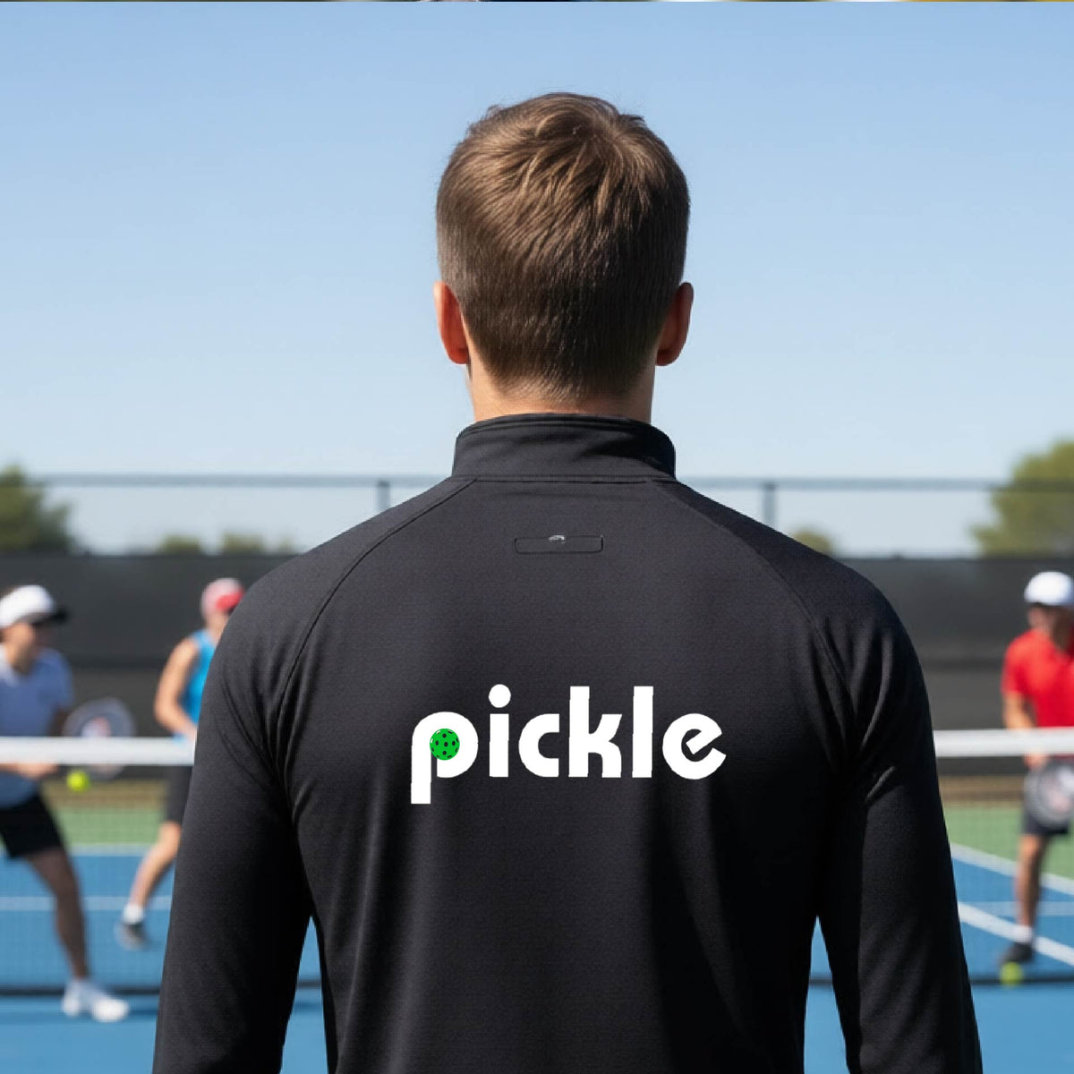 A man wearing a black quarter-zip shirt with a white graphic of ‘PICKLE’ with a green pickleball integrated into the letter P printed on the back in front of a pickleball court. 