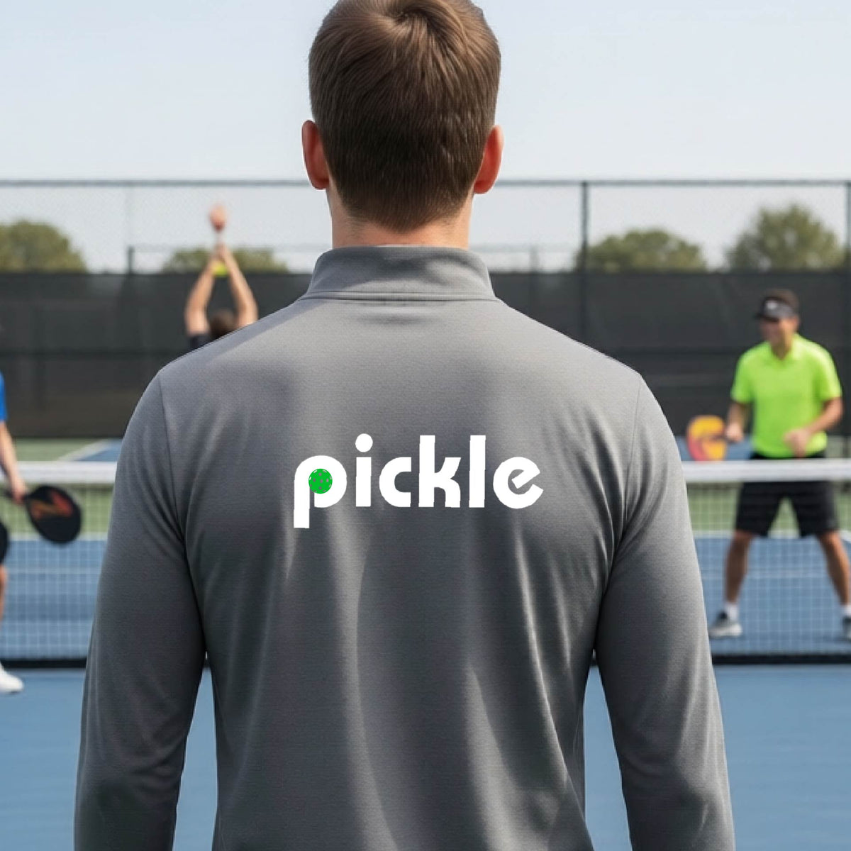 A man wearing a gray quarter-zip shirt with a white graphic of ‘PICKLE’ with a green pickleball integrated into the letter P printed on the back in front of a pickleball court. 
