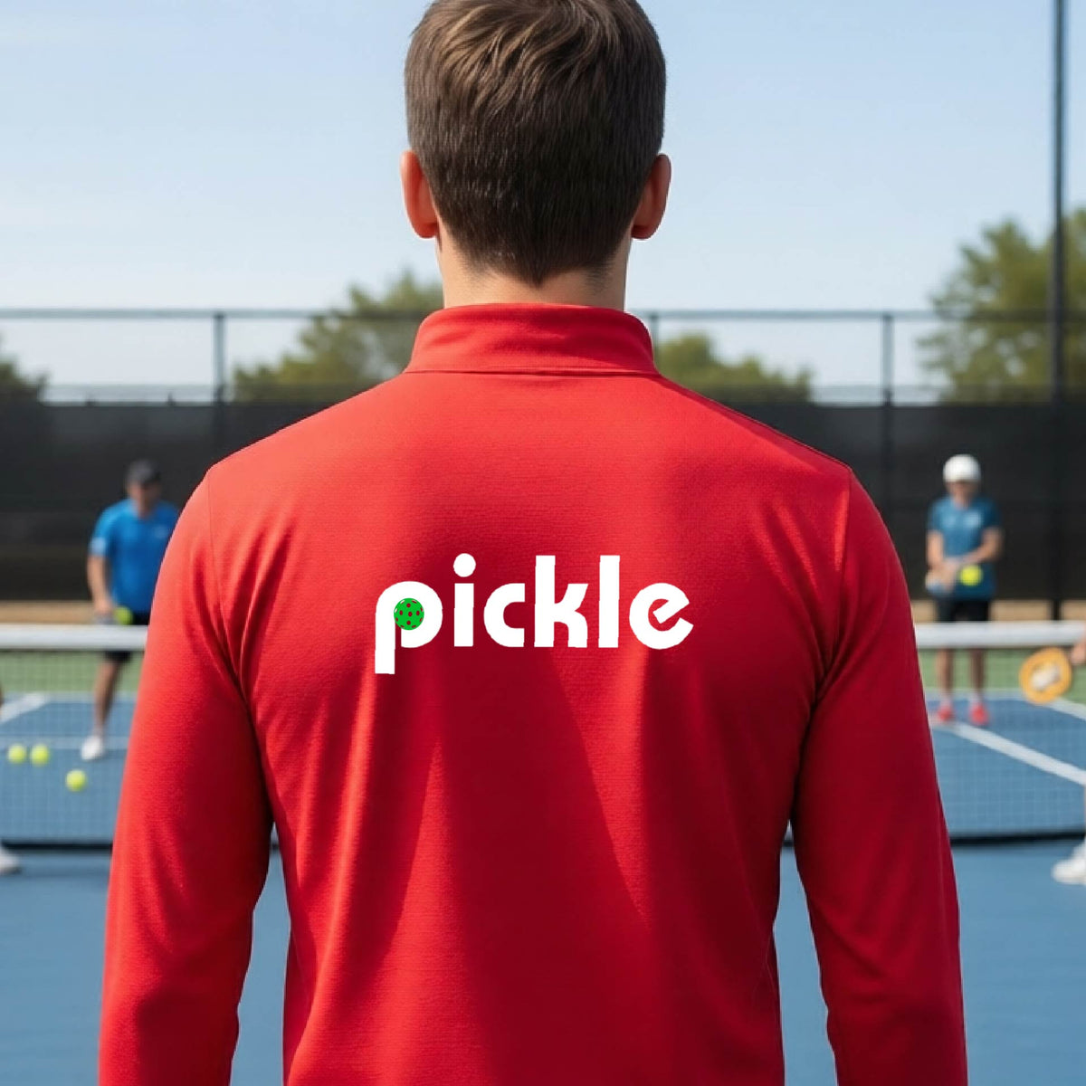 A man wearing a red quarter-zip shirt with a white graphic of ‘PICKLE’ with a green pickleball integrated into the letter P printed on the back, in front of a pickleball court. 