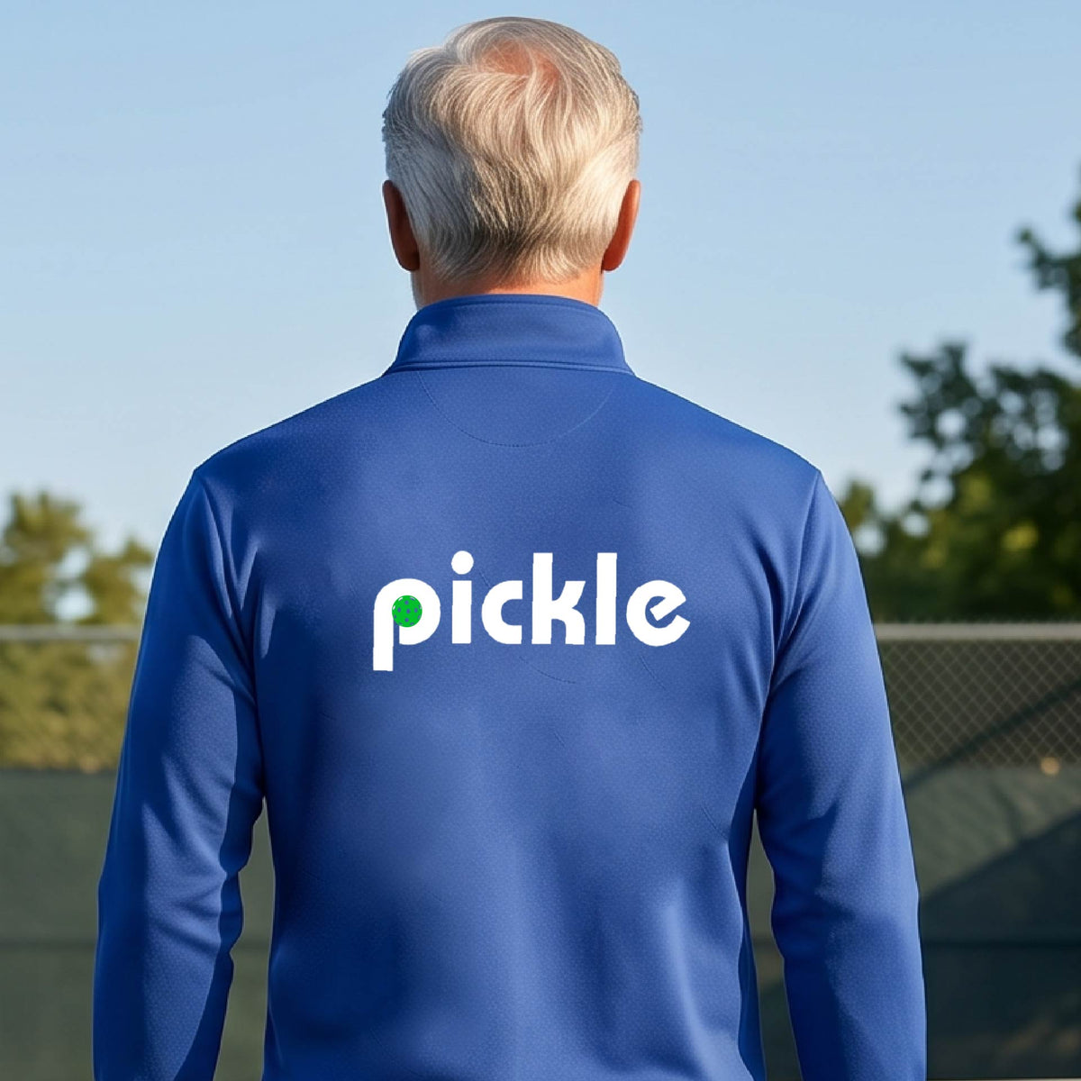 A man wearing a royal blue quarter-zip shirt with a white graphic of ‘PICKLE’ with a green pickleball integrated into the letter P printed on the back in front of a pickleball court. 