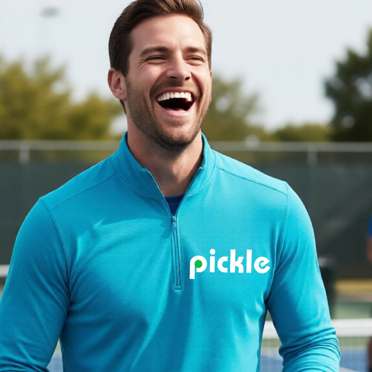 A man wearing a blue quarter-zip shirt with a white graphic of ‘PICKLE’ with a green pickleball integrated into the letter P printed on the front, in front of a pickleball court. 