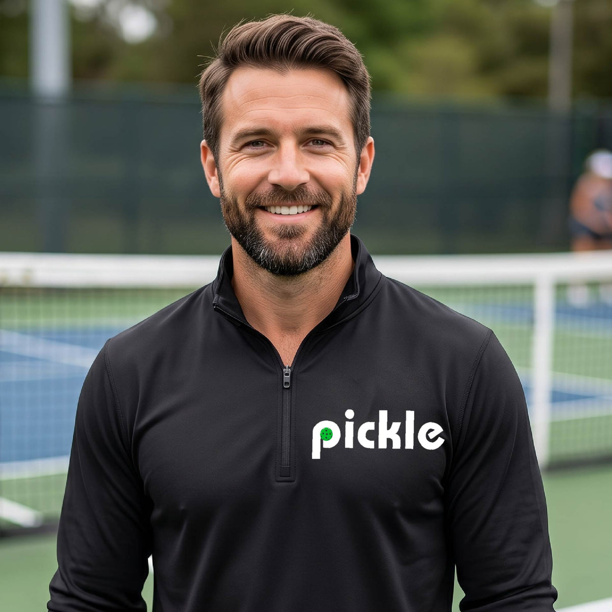A man wearing a black quarter-zip shirt with a white graphic of ‘PICKLE’ with a green pickleball integrated into the letter P printed on the front, in front of a pickleball court. 