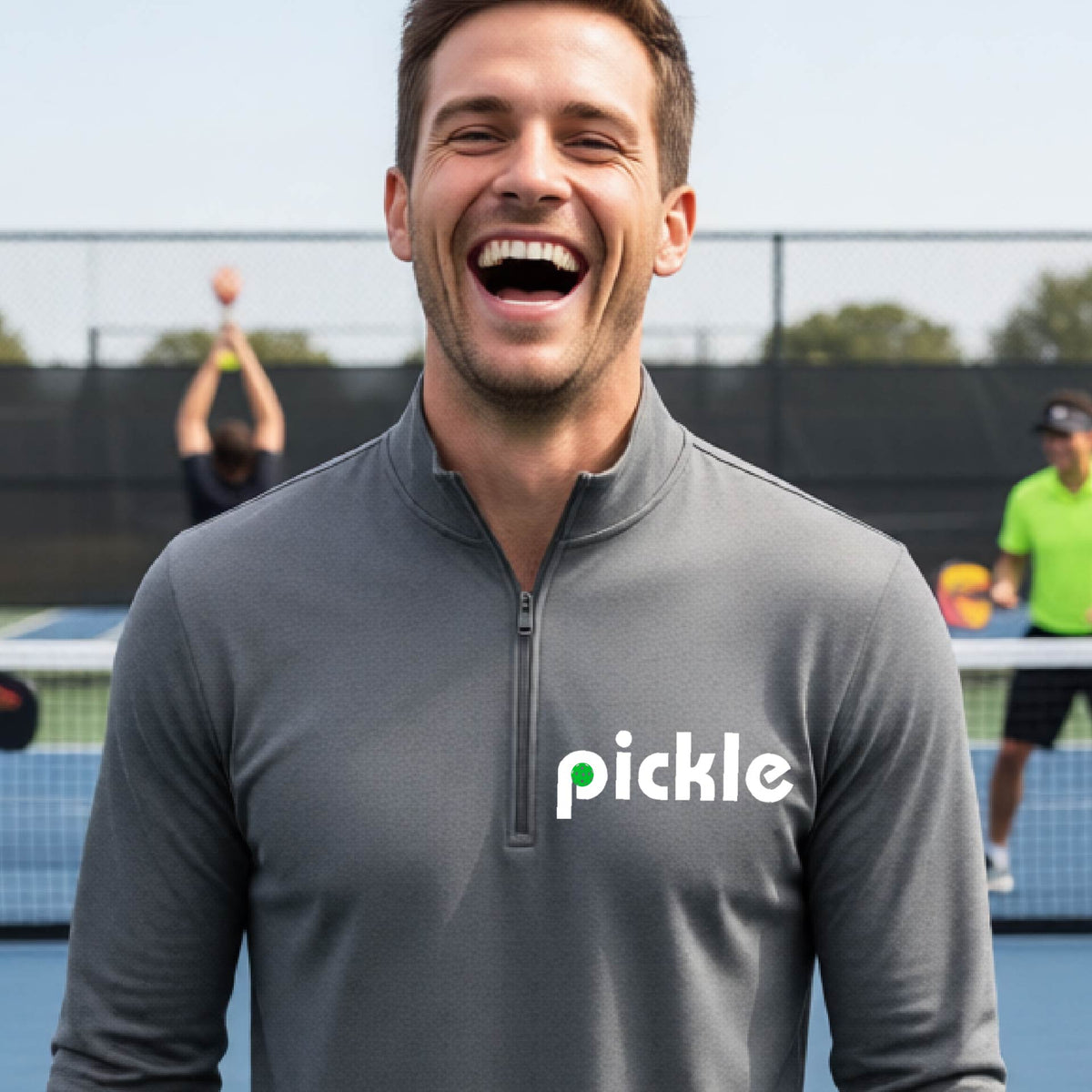 A man wearing a gray quarter-zip shirt with a white graphic of ‘PICKLE’ with a green pickleball integrated into the letter P printed on the front  in front of a pickleball court. 