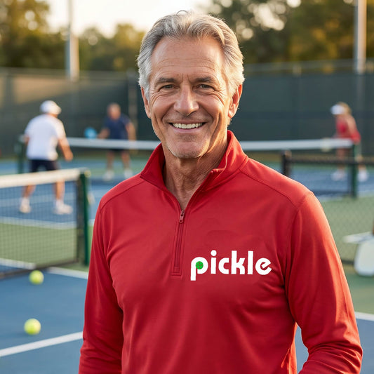 A man wearing a red quarter-zip shirt with a white graphic of ‘PICKLE’ with a green pickleball integrated into the letter P printed on the front, in front of a pickleball court. 