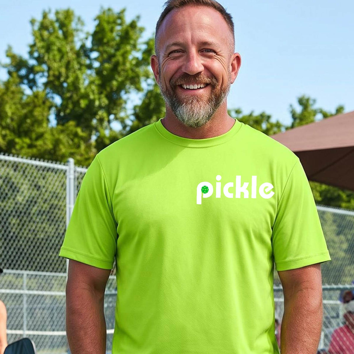 A man wearing a lime green long-sleeve t-shirt with a white graphic of ‘PICKLE’ with a green pickleball integrated into the letter P printed on the front, in front of a pickleball court. 