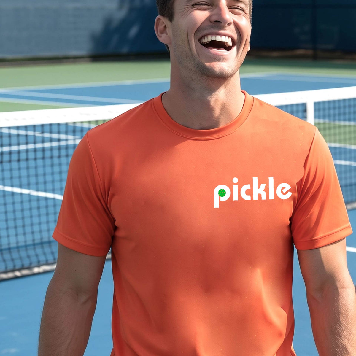A man wearing an orange long-sleeve t-shirt with a white graphic of ‘PICKLE’ with a green pickleball integrated into the letter P printed on the front, in front of a pickleball court. 