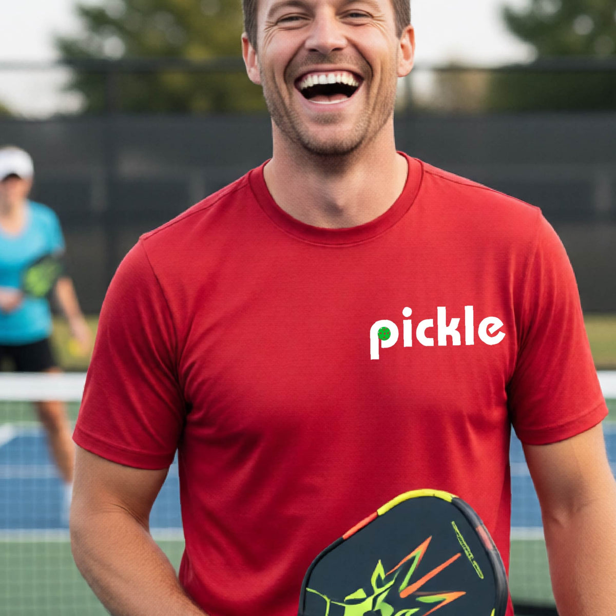 A man wearing a red long-sleeve t-shirt with a white graphic of ‘PICKLE’ with a green pickleball integrated into the letter P printed on the front, in front of a pickleball court. 