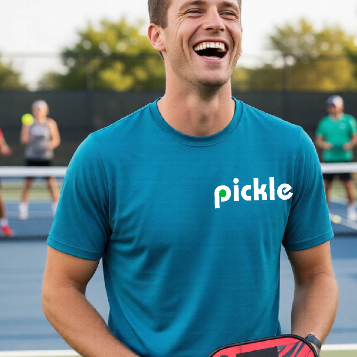 A man wearing a teal long-sleeve t-shirt with a white graphic of ‘PICKLE’ with a green pickleball integrated into the letter P printed on the front, in front of a pickleball court. 