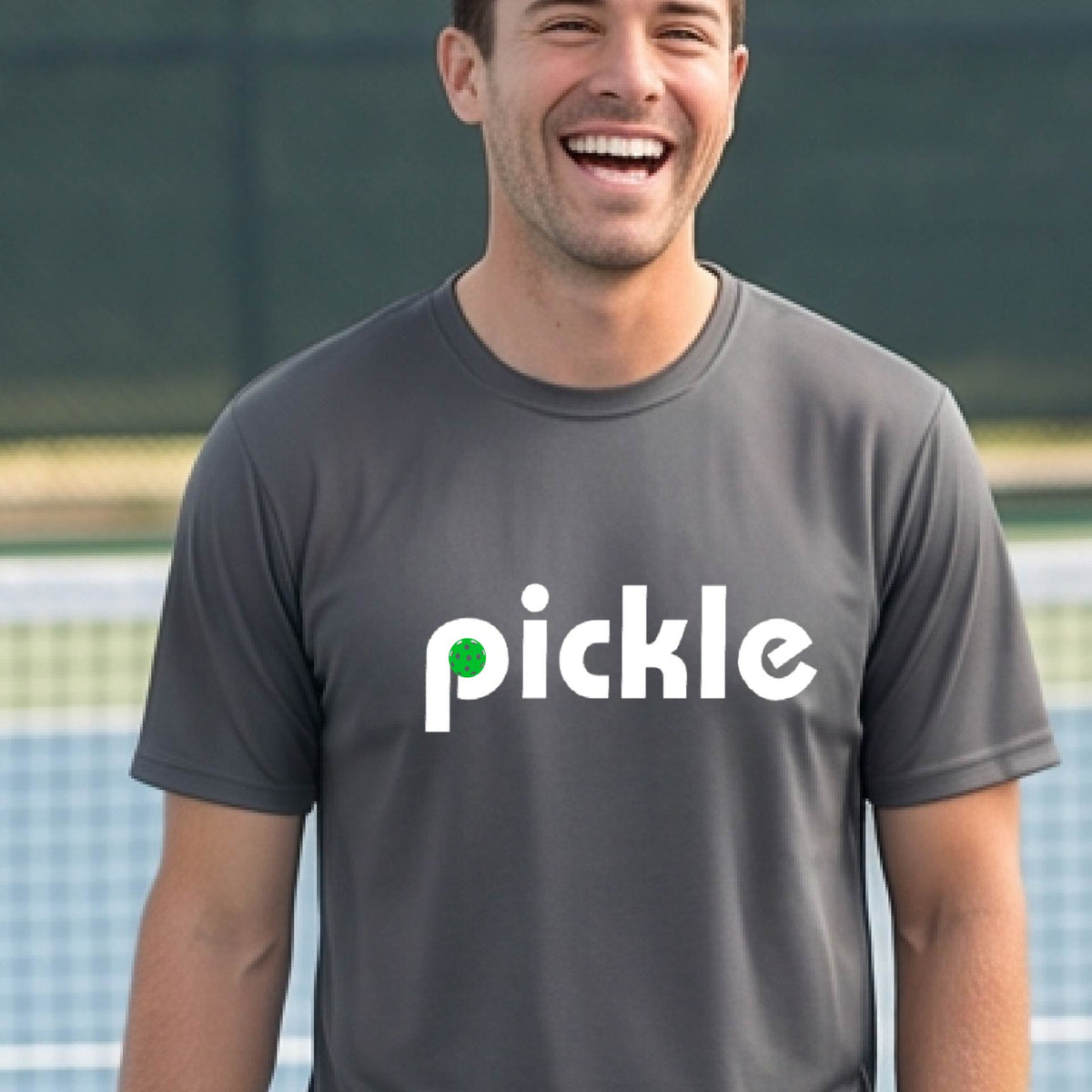 A man wearing a gray long-sleeve t-shirt with a white graphic of ‘PICKLE’ with a green pickleball integrated into the letter P printed on the front, in front of a pickleball court. 