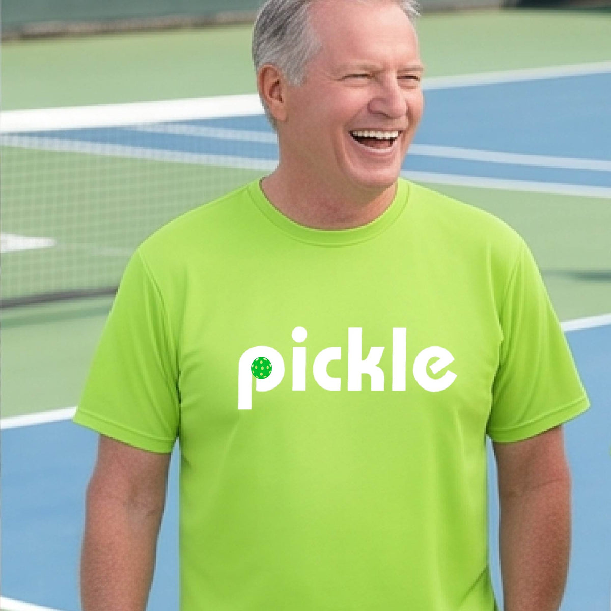 A man wearing a lime green long-sleeve t-shirt with a white graphic of ‘PICKLE’ with a green pickleball integrated into the letter P printed on the front, in front of a pickleball court. 