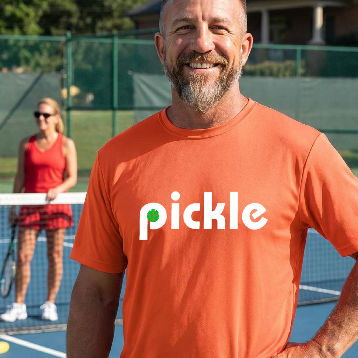 A man wearing an orange long-sleeve t-shirt with a white graphic of ‘PICKLE’ with a green pickleball integrated into the letter P printed on the front, in front of a pickleball court. 
