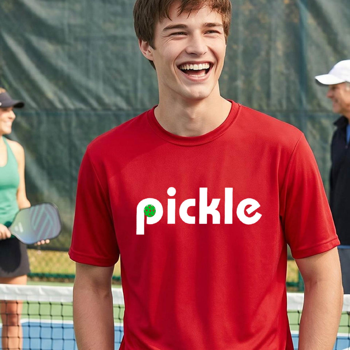 A man wearing a red long-sleeve t-shirt with a white graphic of ‘PICKLE’ with a green pickleball integrated into the letter P printed on the front, in front of a pickleball court. 
