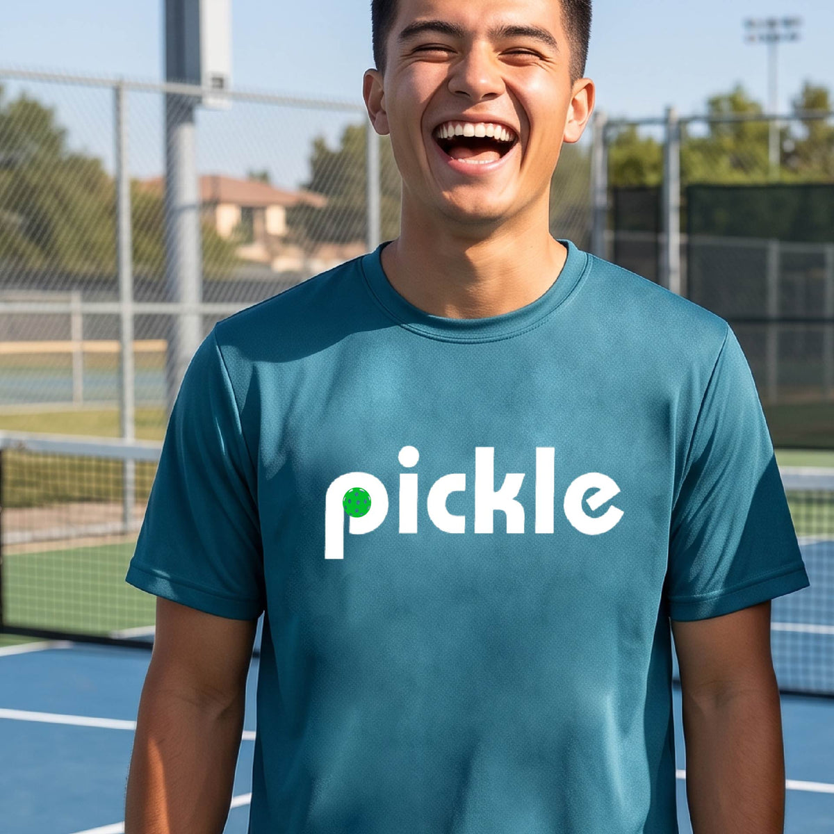 A man wearing a teal long-sleeve t-shirt with a white graphic of ‘PICKLE’ with a green pickleball integrated into the letter P printed on the front, in front of a pickleball court. 