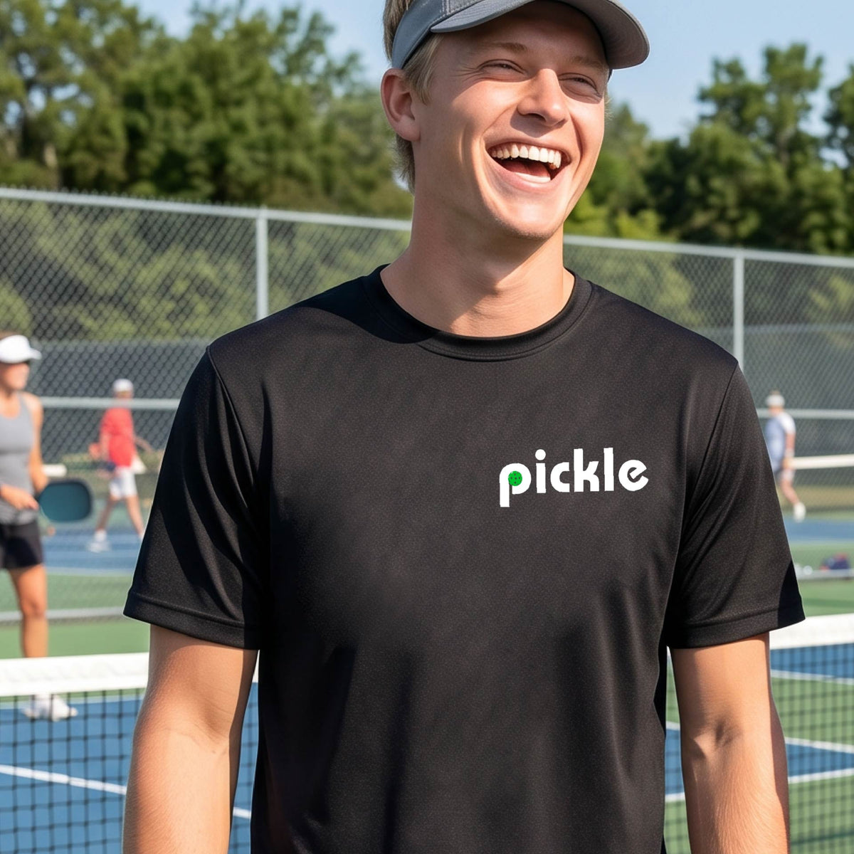 A man wearing a black long-sleeve t-shirt with a white graphic of ‘PICKLE’ with a green pickleball integrated into the letter P printed on the front, in front of a pickleball court. 