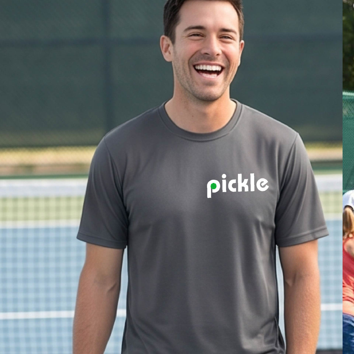 A man wearing a gray long-sleeve t-shirt with a white graphic of ‘PICKLE’ with a green pickleball integrated into the letter P printed on the front, in front of a pickleball court. 