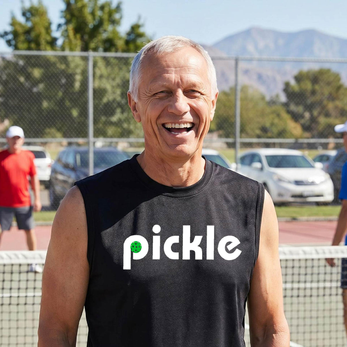 A man wearing a black sleeveless t-shirt with a white graphic of ‘PICKLE’ with a green pickleball integrated into the letter P printed on the front, in front of a pickleball court. 