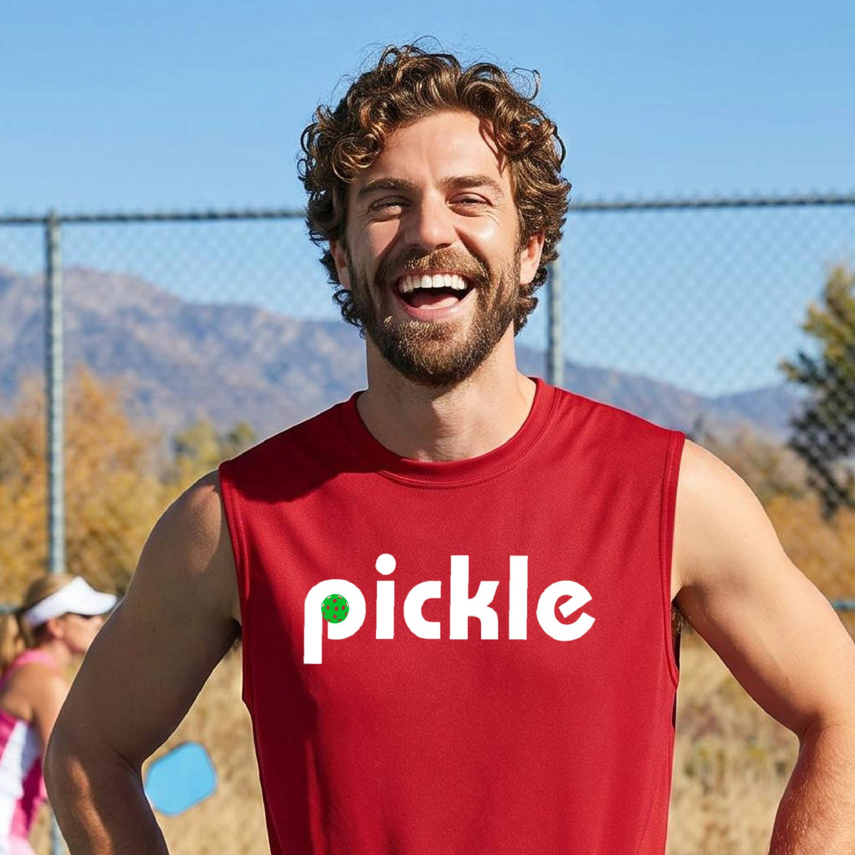 A man wearing a red sleeveless t-shirt with a white graphic of ‘PICKLE’ with a green pickleball integrated into the letter P printed on the front, in front of a pickleball court. 