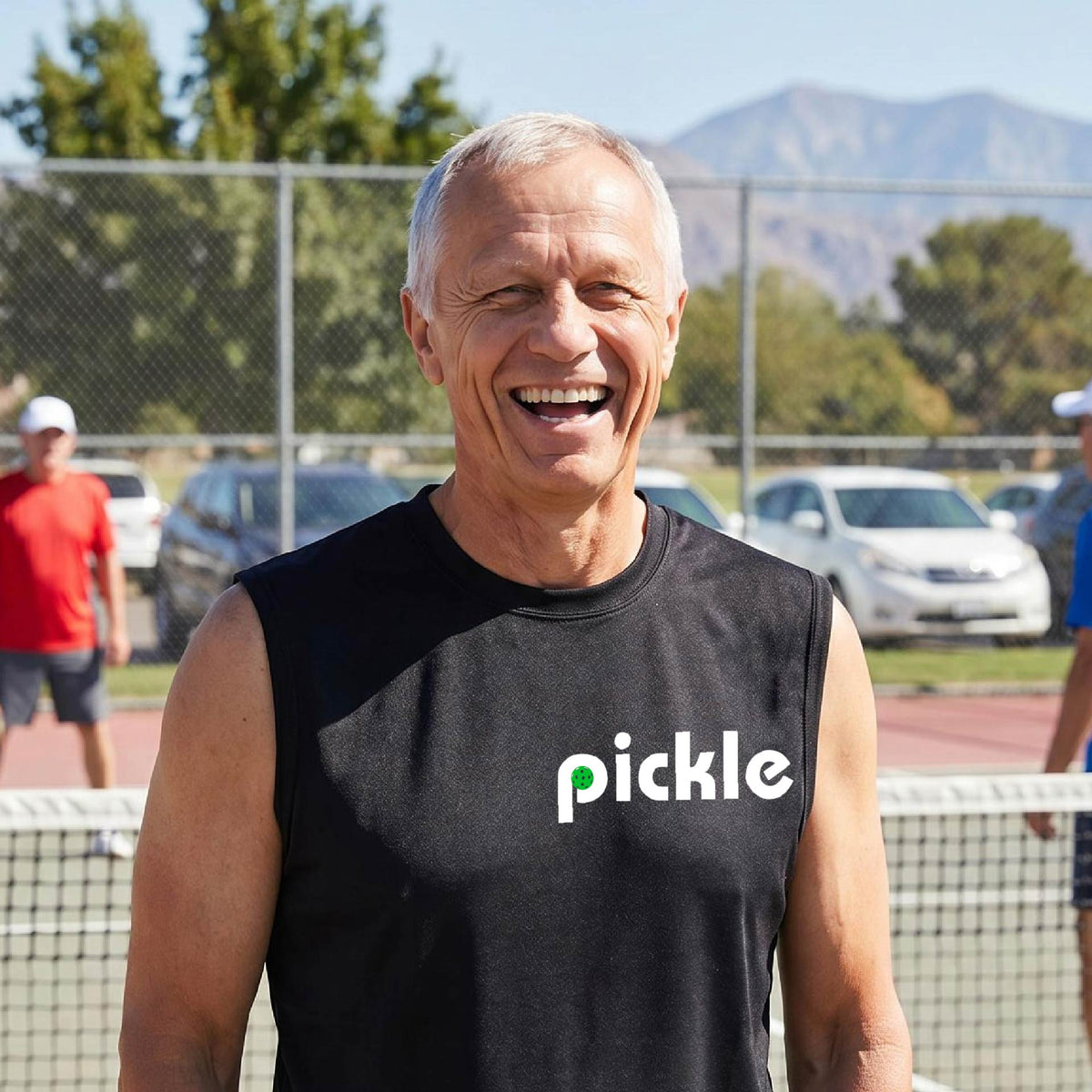 A man wearing a black sleeveless t-shirt with a white graphic of ‘PICKLE’ with a green pickleball integrated into the letter P printed on the front, in front of a pickleball court. 