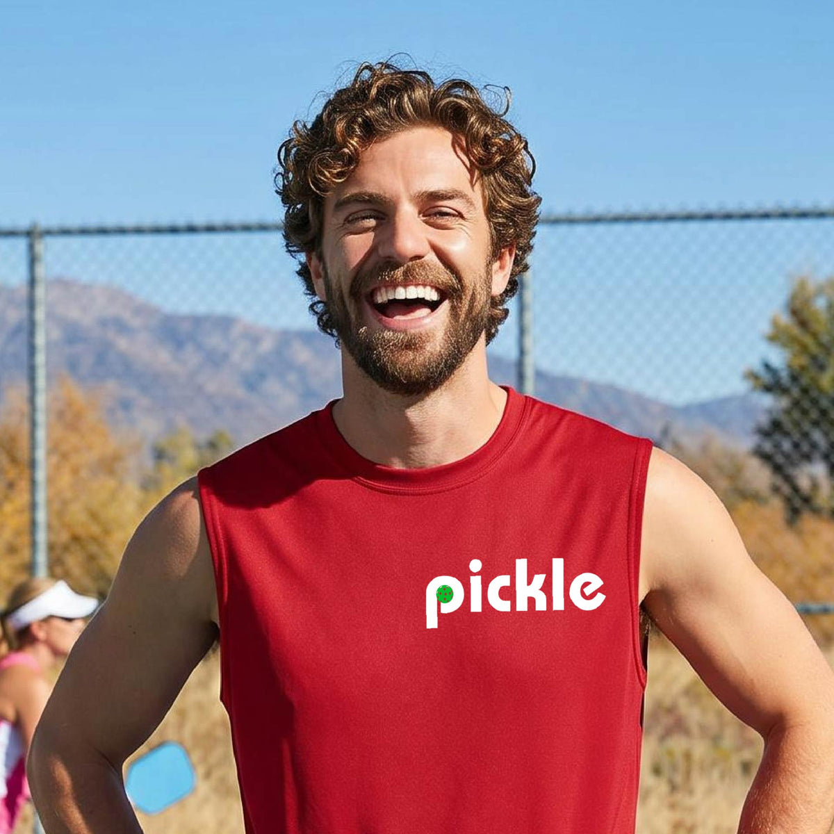 A man wearing a red sleeveless t-shirt with a white graphic of ‘PICKLE’ with a green pickleball integrated into the letter P printed on the front, in front of a pickleball court. 
