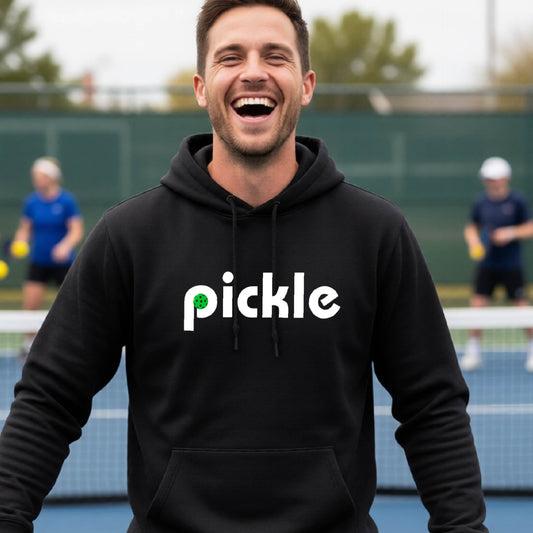A close-up of a black unisex hoodie pullover with a white graphic of ‘PICKLE’ with a green pickleball integrated into the letter P printed on the front, in front of a pickleball court. 
