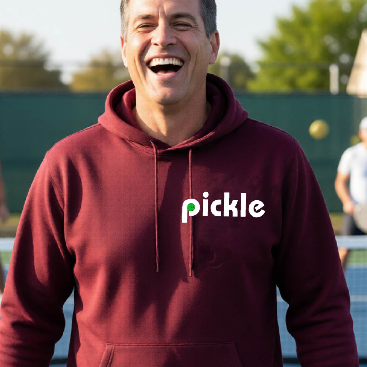 A close-up of a maroon unisex hoodie pullover with a white graphic of ‘PICKLE’ with a green pickleball integrated into the letter P printed on the front, in front of a pickleball court. 