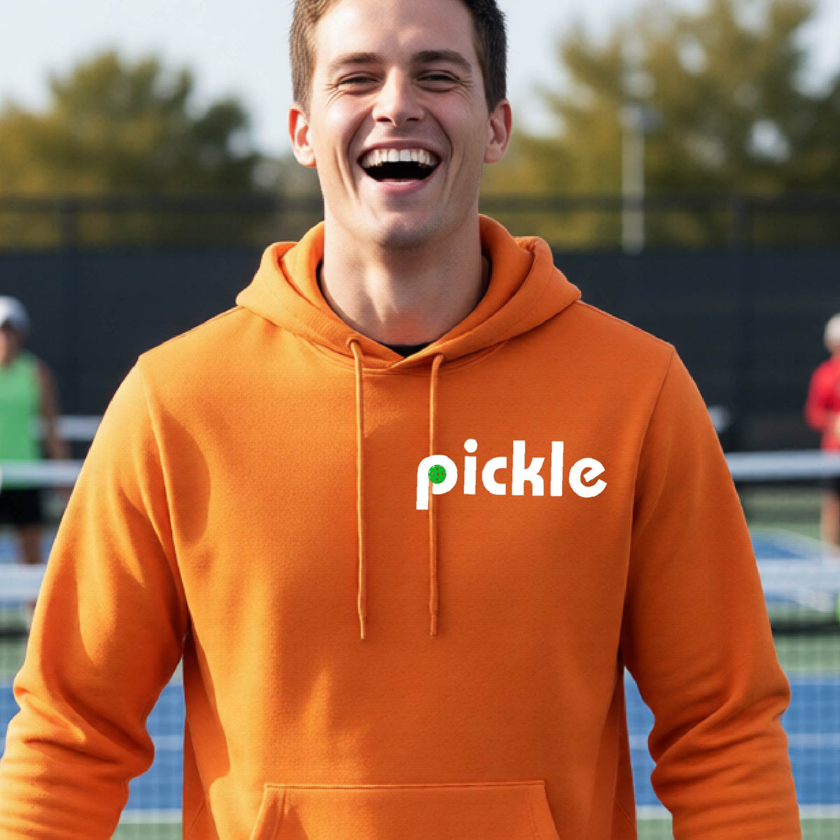 A close-up of n orange unisex hoodie pullover with a white graphic of ‘PICKLE’ with a green pickleball integrated into the letter P printed on the front, in front of a pickleball court. 