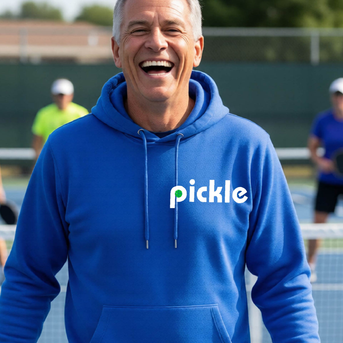 A close-up of a royal blue unisex hoodie pullover with a white graphic of ‘PICKLE’ with a green pickleball integrated into the letter P printed on the front, in front of a pickleball court. 