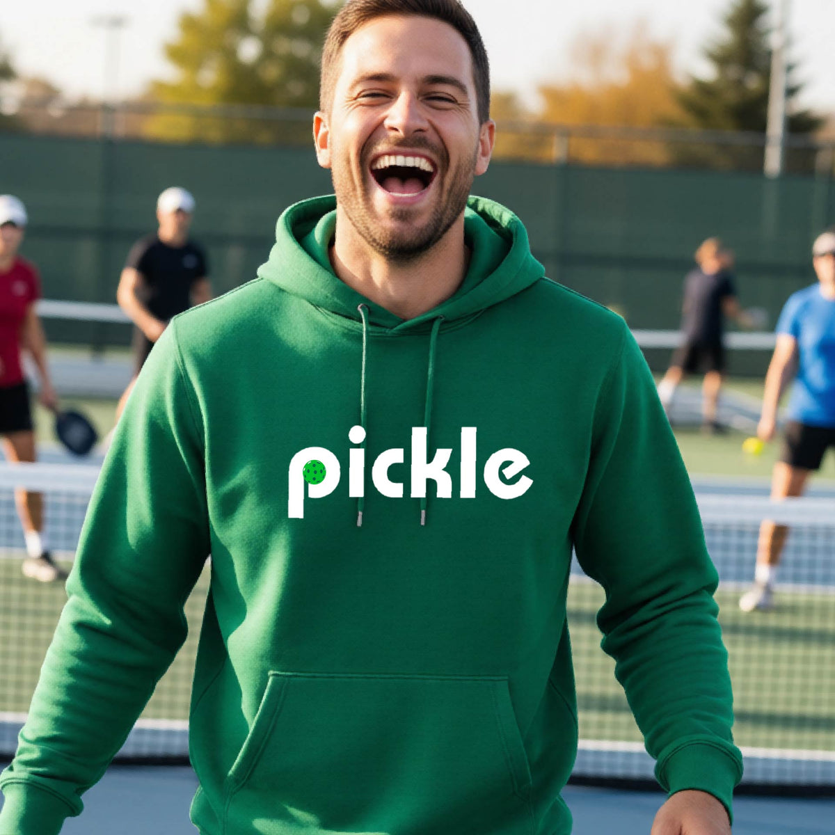 A close-up of a green unisex hoodie pullover with a white graphic of ‘PICKLE’ with a green pickleball integrated into the letter P printed on the front, in front of a pickleball court. 