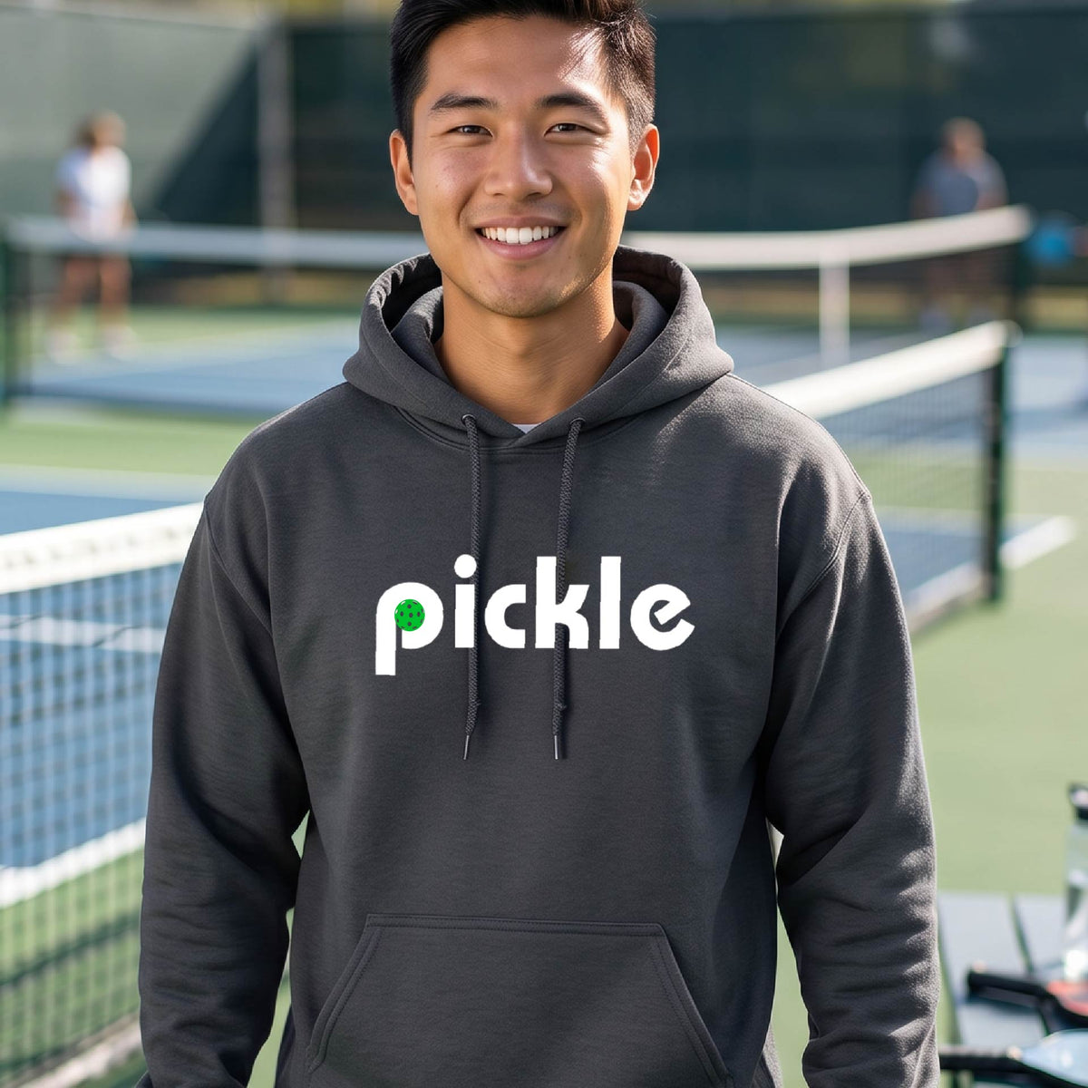 A close-up of a gray unisex hoodie pullover with a white graphic of ‘PICKLE’ with a green pickleball integrated into the letter P printed on the front, in front of a pickleball court. 