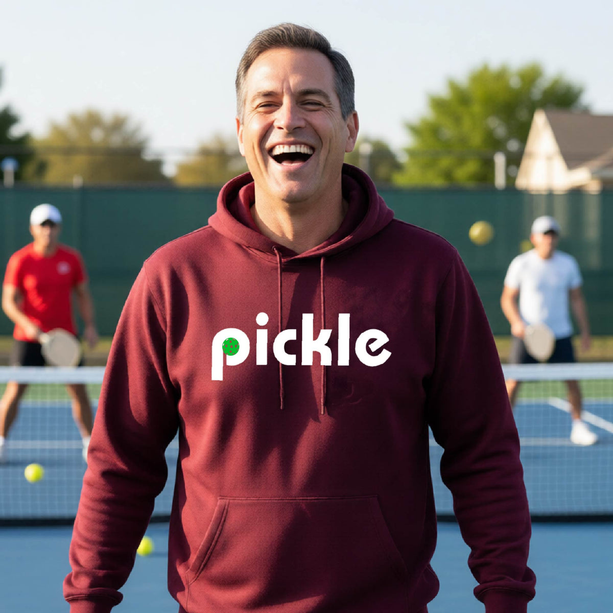 A close-up of a maroon unisex hoodie pullover with a white graphic of ‘PICKLE’ with a green pickleball integrated into the letter P printed on the front, in front of a pickleball court. 