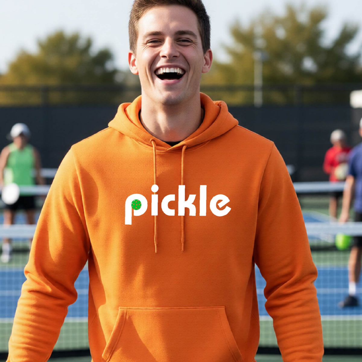 A close-up of n orange unisex hoodie pullover with a white graphic of ‘PICKLE’ with a green pickleball integrated into the letter P printed on the front, in front of a pickleball court. 