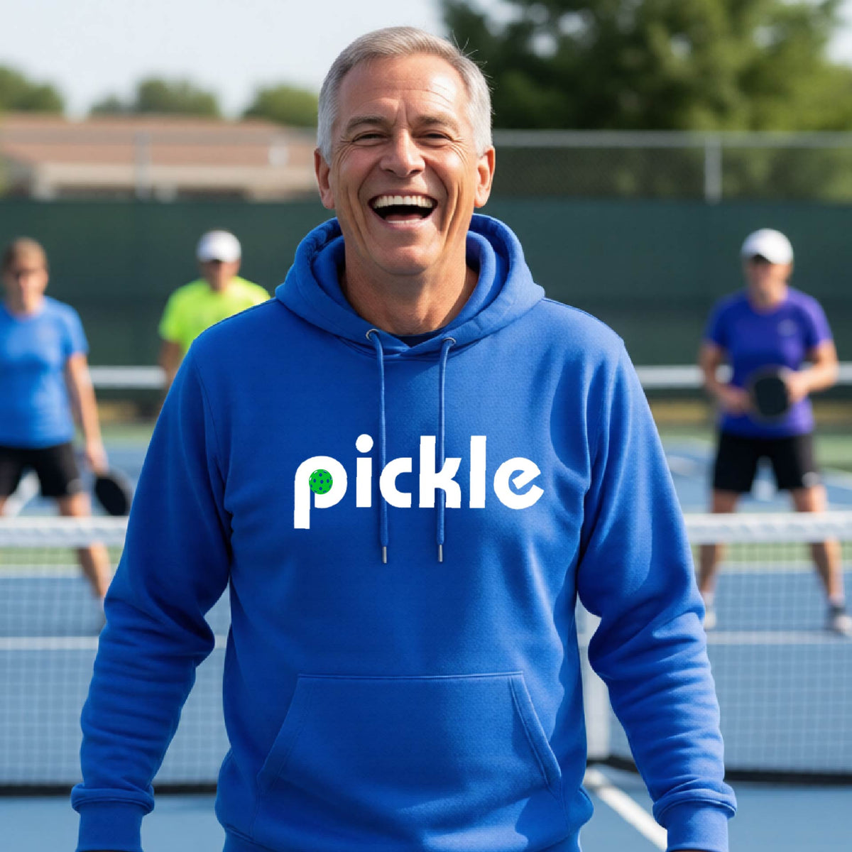 A close-up of a royal blue unisex hoodie pullover with a white graphic of ‘PICKLE’ with a green pickleball integrated into the letter P printed on the front, in front of a pickleball court. 
